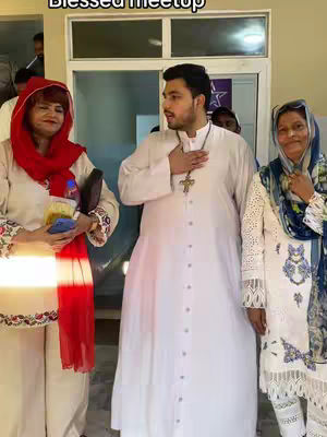 The image shows three people standing indoors. The man in the center is wearing a white robe and a cross necklace, with his hand over his chest. To his left is a woman wearing a red scarf over her head and holding a bottle. To the right is a woman in a white dress. The background includes a door, and the text overlay 'Blessed Meetup'.