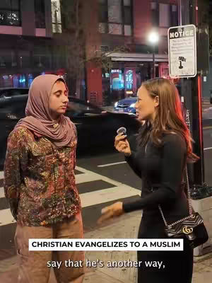 Two women are standing on a city street, conversing. One wears a hijab and a printed jacket, the other is in a black outfit. A sign is partially visible in the background.