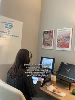 A person sits at a desk in front of a laptop, a tablet, and a glass of beer. There is a paper with text on it. On the wall are two framed art prints, and a whiteboard with notes.