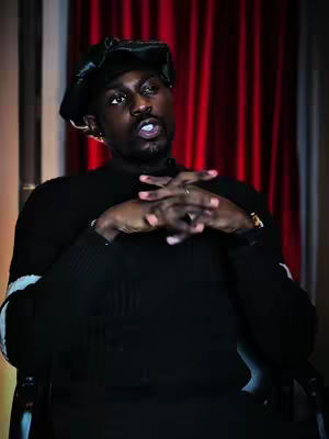 A man in a black sweater and a black hat is seated, looking up and to the right with his hands clasped together, fingers intertwined. The backdrop is a deep red curtain.