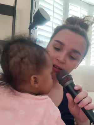 A woman holds a baby close, both seemingly engrossed in holding a microphone. The woman has her hair up, and the baby is facing her. The image is well-lit, and the background shows a window with shuttered blinds.