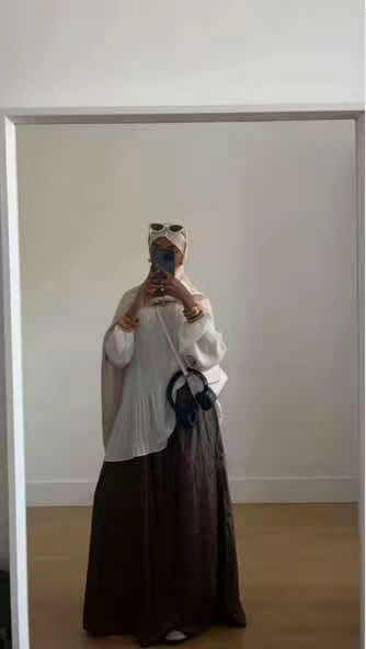 A full-length mirror selfie. The creator is seen from the chest up, wearing a white hijab, a white top, and a long brown skirt. A crossbody bag and headphones are visible. The background is a light-colored wall and a wooden floor.
