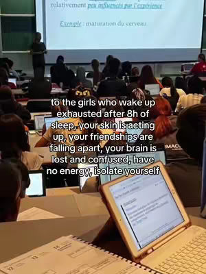 The image captures an audience in a lecture hall, with a large screen displaying text and a presenter in the background. In the foreground, there are multiple tablets, laptops, and papers, indicating a study or academic setting. The overall composition suggests a classroom environment.