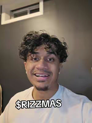 A man with curly hair and a white t-shirt is smiling at the camera. The background is a dark grey wall. The text overlay '$RIZZMAS' in a white font with a black outline is at the bottom.