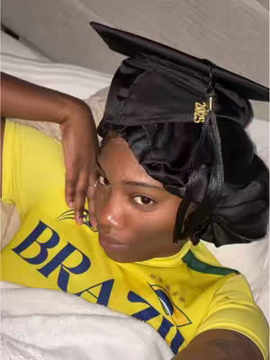 A woman in a yellow shirt and black graduation cap is lying in bed. In the background, the bed has white sheets. The woman's face displays a neutral expression.