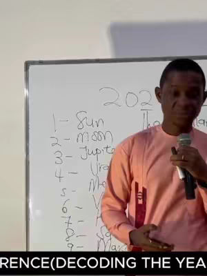 A person is standing in front of a whiteboard, holding a microphone. The whiteboard has the year 202 on top with a list of planets next to numbers underneath. The person is wearing a pink shirt. The camera is angled towards the person.