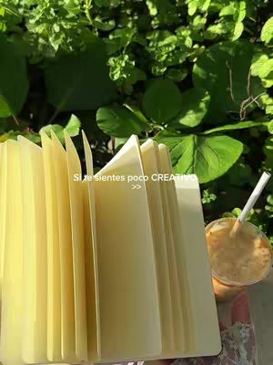 A close-up shot of an open notebook with yellow pages, sitting in front of a blurred background of green foliage and a drink with a straw. The book is the central element. The drink is on the right side.