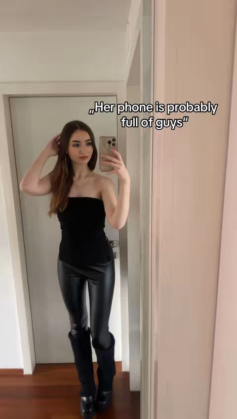 A woman in a black tube top, black leather pants, and black boots stands in a mirror, taking a selfie with her phone. Text is overlaid near the top of the image.