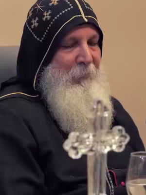 A close-up shot features a religious figure. He's an older man, with a long white beard and wearing a black hooded garment. The hood is detailed with embroidered patterns. A clear cross is in the foreground. The composition is focused on the individual, suggesting a devotional or contemplative theme, with a muted background that is out of focus.