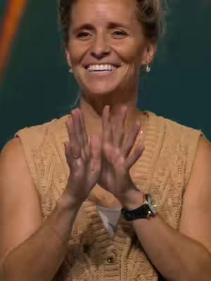 A woman with fair skin, blonde hair pulled back, and wearing a beige sweater vest smiles broadly, clapping her hands together. The background features a blurred green.