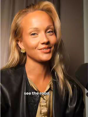 A woman with blonde hair and a black leather jacket is looking toward the camera and smiling. Her make up includes blush and lip color. She also has small gold earrings. Text overlayed says 'see the road'.