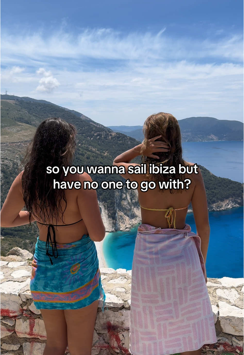 Two women, viewed from the back, stand on a stone ledge overlooking a scenic coastal view. The woman on the left wears a black bikini top and a blue patterned wrap. The woman on the right wears a yellow bikini top and a pink wrap. The background features a vast turquoise sea, a white sand beach, and mountainous terrain under a bright blue sky with wispy clouds.