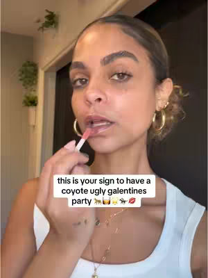 A woman with dark hair and light skin is applying lip gloss. She has gold hoop earrings and a white top. A text overlay in a white box reads "this is your sign to have a coyote ugly galentines party" with various related emojis.