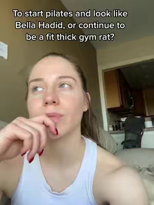 A woman with light skin and light brown hair tied back in a ponytail is shown in a selfie-style shot. She is wearing a white tank top and has red nail polish. She is holding her hand up to her face, with her fingers near her lips as if in thought or considering an idea. In the background, a kitchen area is partially visible, with wooden cabinets and a glimpse of the room, adding to the casual, everyday setting of the video.