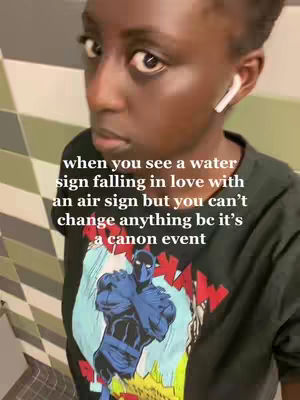 A selfie-style shot with the creator looking at the camera. The background appears to be a tiled wall. Text overlay on the image reads, 'when you see a water sign falling in love with an air sign but you can't change anything bc it's a canon event'. The creator is wearing a graphic t-shirt with a design centered on the chest. They have one white earbud visible in ear.