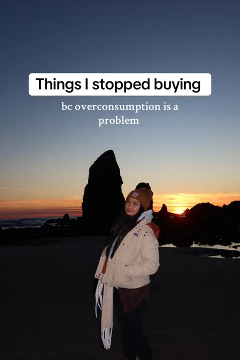 A woman stands on a beach at sunset, the sun casting an orange glow over the water and silhouetting distant rock formations. Text overlays read 'Things I stopped buying' and 'bc overconsumption is a problem.