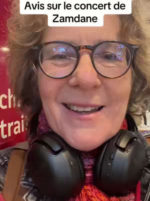 A selfie-style video with a woman wearing glasses and headphones. The background is a blurred interior scene.