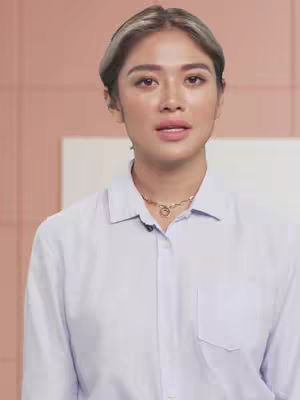 A woman with light skin, blonde-brown hair and a light blue button up shirt stands facing the camera. The background consists of a pale pink wall and an off-white space on the left side. She is wearing a gold necklace and her expression is neutral.