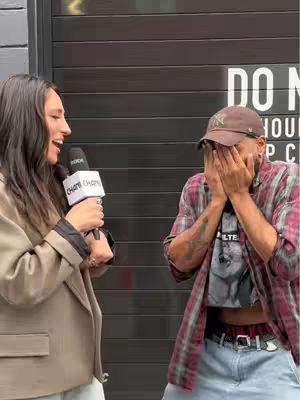 A woman with dark hair is holding a microphone and looking to the right. A man wearing a baseball cap is covering his face with his hands. They are standing outside against a dark background.