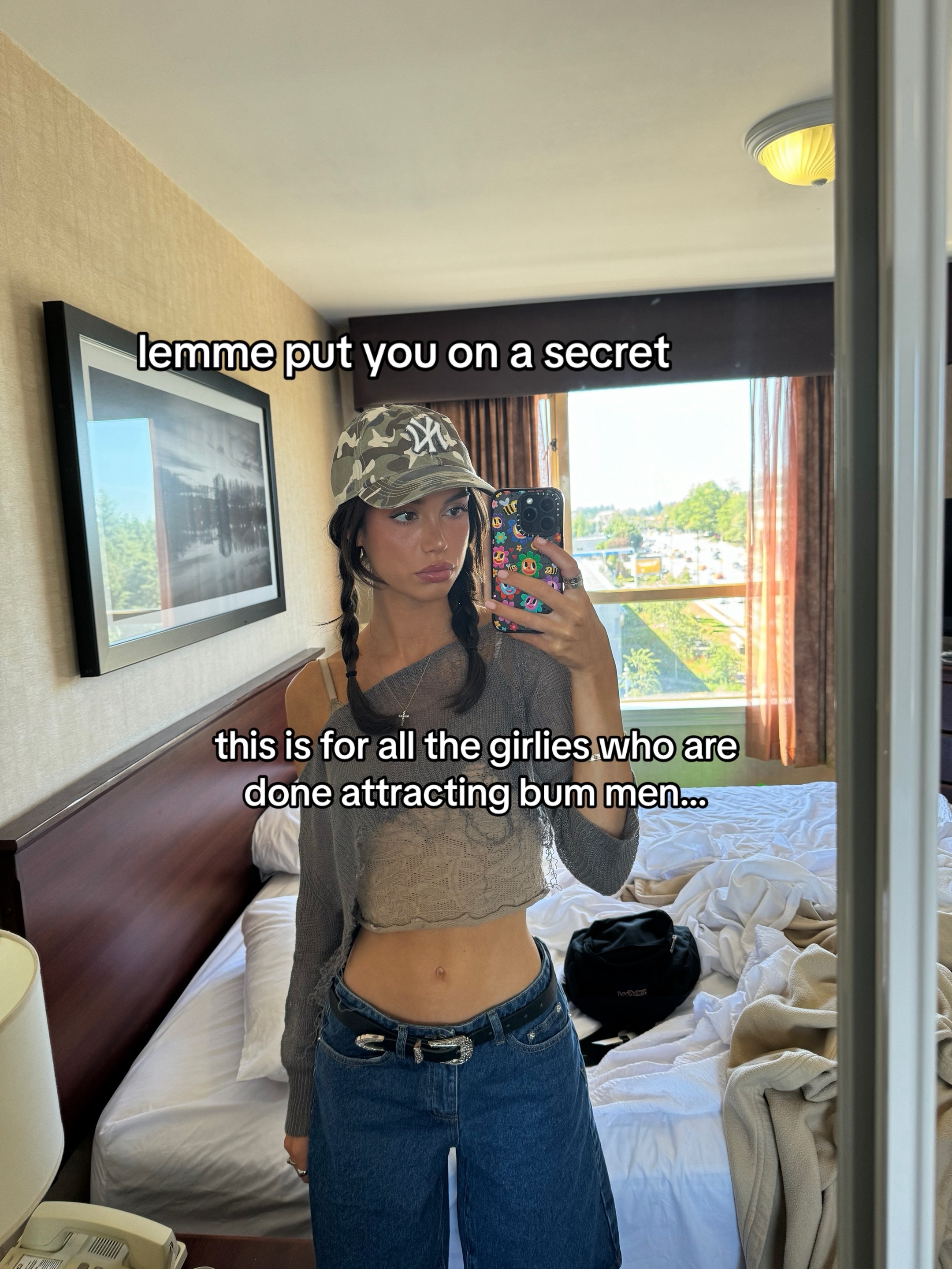 A woman is taking a selfie in her hotel room. She's wearing a camouflage baseball cap, a crop top, and low-rise jeans, and she's holding her phone up to take the picture. Behind her is a large bed with a patterned bedspread and a mirror that reflects the room's surroundings.