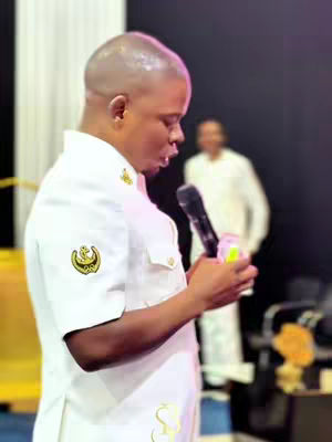 A man in a white uniform with gold accents, holding a microphone and speaking. Another person in white is blurred in the background. The lighting suggests an indoor setting, possibly a church or event. The overall composition is centered around the speaker.