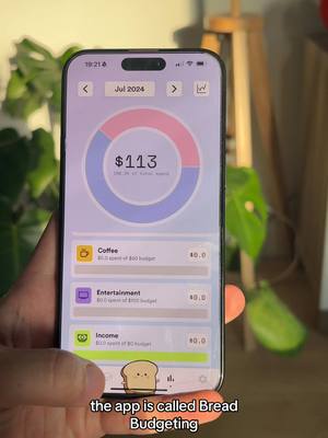 A person's hand holding a smartphone displaying an app interface. The phone screen showcases a budgeting app with a circular progress chart displaying $113. Below, there are sections for 'Coffee,' 'Entertainment,' and 'Income,' with associated spending details. A whimsical bread character appears at the bottom. The background includes a blurry green plant and a wooden frame.