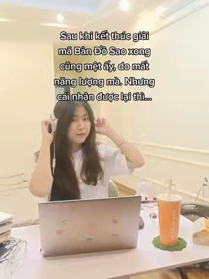 A woman is sitting at a desk with a laptop and a drink. She is looking at the camera while wearing headphones.