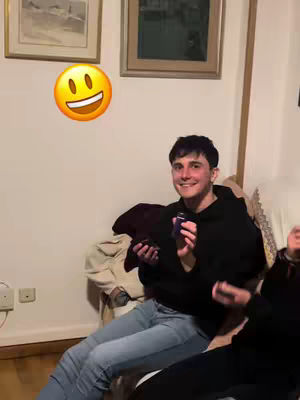 A man in a black hoodie and jeans sits on a sofa, smiling and holding a small purple object, possibly a card or bottle. An emoji hovers above him. The setting appears to be an indoor space, perhaps a living room, with some light colors and simple wall decor and artwork.