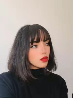 A young woman with shoulder-length dark hair and bangs is the focus of the picture. She looks off to her right with a red lip. She's wearing a black turtleneck and the background is a plain wall.