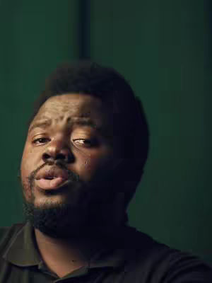 Close-up shot of a Black man with a short beard and dark hair. He appears to be emotional, with a tear visible on his cheek and his lips slightly parted as if he is about to speak. The background is a solid, muted green.