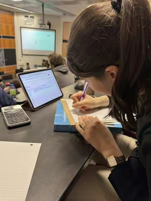 A classroom setting with a student in the foreground. The student, with dark hair pulled up in a ponytail, is seated at a desk, writing on a pad of paper with a pen. An open laptop and a calculator sit to the left of the student's writing materials. Behind the student, a whiteboard and a large projection screen are visible, indicating a lecture or presentation.