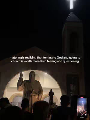 The image shows a nighttime outdoor church scene. A large, illuminated statue is visible inside a rounded archway. In front of the church, a crowd of people is gathered, some holding up phones. A text overlay provides additional context.
