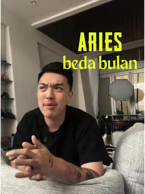 A man with tattoos on his arm is seated on a couch. The hook says ARIES beda bulan with a confused expression.