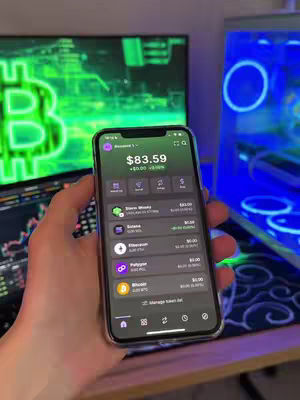 A hand holding a smartphone displaying a cryptocurrency portfolio, with a blurred background showing a computer monitor and other tech items.