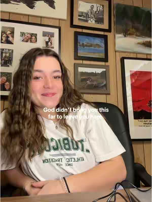 A young woman with long brown hair sits in front of a desk, smiling at the camera. Behind her are various framed pictures on a wall. The lighting is natural, and she's wearing a white t-shirt.