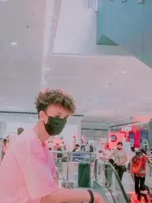 A selfie-style shot of a young man at a mall. He's wearing a pink shirt and black face mask. The background shows a mall environment with people, escalators, and other details.