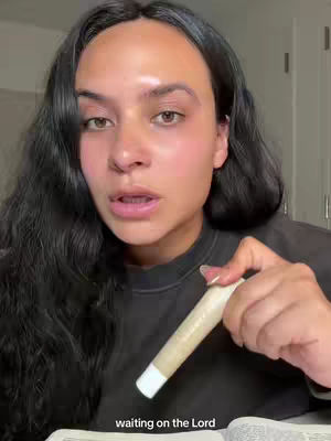 A woman with long, dark hair is holding a rolled paper item in her right hand. She has fair skin and is looking directly at the camera with a slight expression. The background is slightly blurred, suggesting an interior setting with neutral tones.