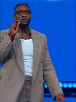 A medium shot features a Black man, dressed in a tan pinstriped suit, gesturing with his right hand as he speaks. The suit has a double-breasted design, and he wears a white undershirt beneath. His glasses and a silver necklace are visible. The background is a solid, bright blue.