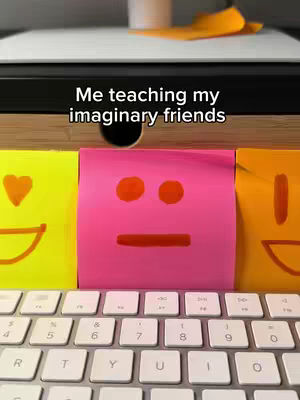 The image shows three colorful sticky notes with drawn faces, placed above a white keyboard with the keyboard in the foreground and a wooden structure.