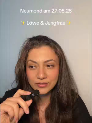 A woman with brown hair holds a small black microphone, looking slightly to her left. Over her head, text reads: "Neumond am 27.05.25" and "✨ Löwe & Jungfrau ✨". The background is a plain wall.
