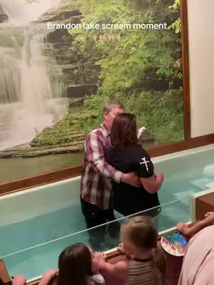 A man and woman are embracing in a baptismal font, with others in the background. Behind them is a large mural of a waterfall and greenery, indicating a church setting.
