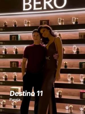 The cover image shows a couple posed in front of a display shelf, likely in a store. The man is on the left, wearing a maroon shirt and black pants, with his hands in his pockets. The woman is taller, wearing a long, dark dress, and has her arm around his shoulder. Text overlayed at the bottom reads 'Destino 11 Pareja.' The background consists of shelves filled with products. The overall color palette is neutral, with the focus on the couple.