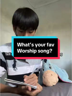 A person is seen looking down at a notebook while holding a pen. In the background, there is a stuffed donkey toy and a text overlay that reads, "What's your fav Worship song?" The overall composition is casual and appears to be an intimate setting.