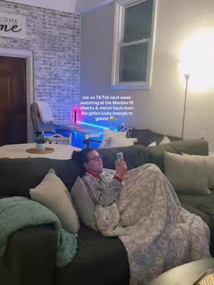 A woman is seated on a dark green couch, wrapped in a patterned blanket, holding a phone. The room features a white brick wall on the left and a window on the right, with a floor lamp casting a soft glow. Text overlay is visible.