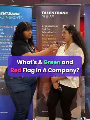 The image shows two women in an interview setting, with banners in the background. A large purple overlay with white text stating "What's A Green and Red Flag In A Company?" is superimposed at the bottom. The woman on the left is wearing a black top and jeans, while the woman on the right is wearing a white top and black pants. Both are looking towards each other, and the woman on the right is holding a microphone towards the other.