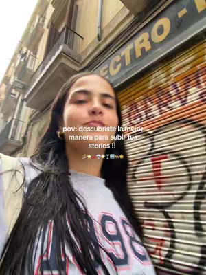 A selfie-style shot featuring a woman with long, dark hair standing outdoors. She is positioned slightly off-center against a backdrop of a building facade with a weathered appearance. The background also includes a section of a shop shutter with graffiti. The woman is wearing a grey t-shirt with graphic elements and has gold hoop earrings. Text overlays the image near the top, with a series of small emojis. The lighting appears to be natural daylight, with a slight blur.