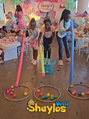 A group of young people are playing a game at a birthday party. They each have a pole with a hoop at the end and are attempting to catch balls suspended in mid-air. There is a birthday banner in the background, and balloons.