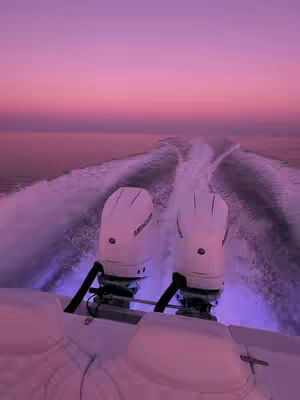 A view from the back of a speeding boat at sunset, with the wake trailing behind. Two white boat engines, with "Mercury" logos, are the primary focus, framed by the white boat seats in the foreground. The sky is a gradient of pink and purple, and the water reflects these colors.