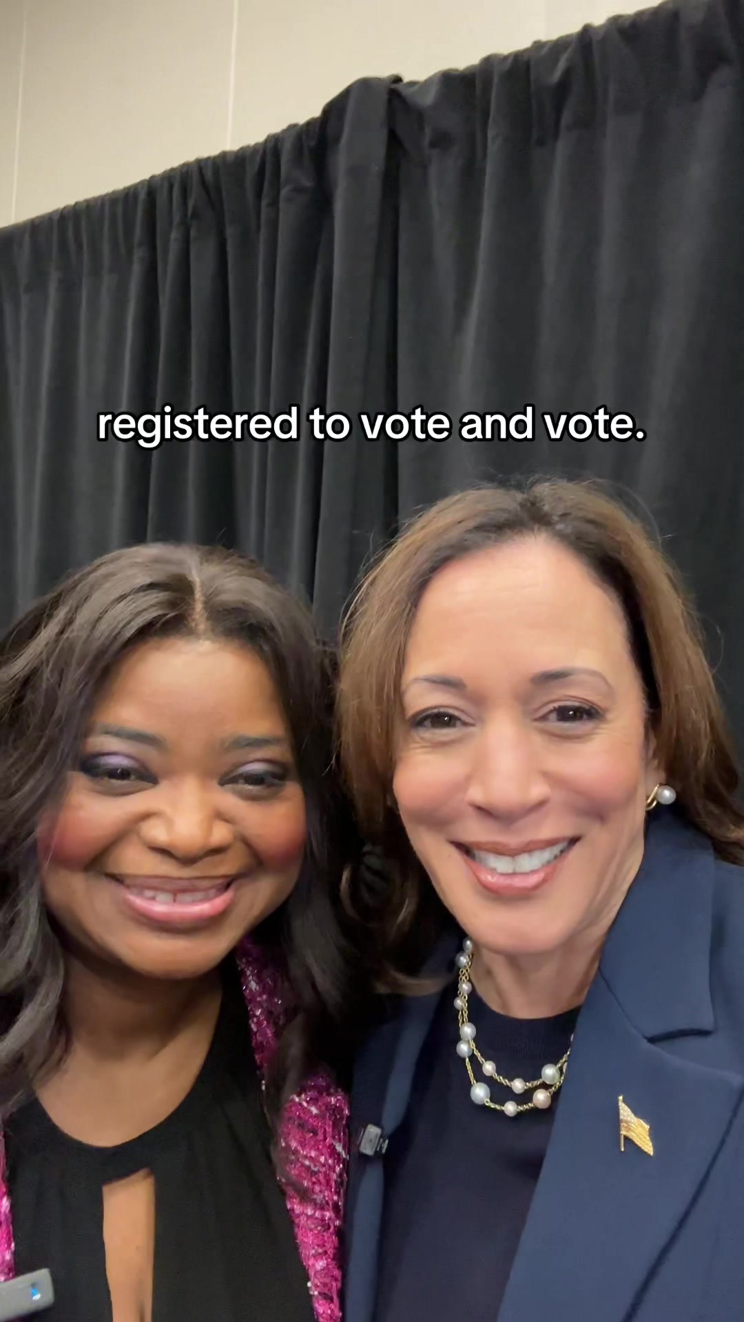 Two smiling women are captured in a selfie-style shot. The woman on the left has dark hair and is wearing a pink jacket. She is smiling at the camera. The woman on the right has brown hair and a navy blue blazer. They are standing in front of a dark curtain.