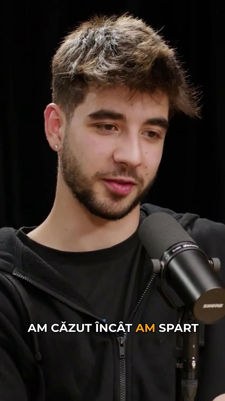 A medium shot features a young man, the primary focus, set against a plain, dark background. He is fair-skinned with short, light brown hair styled up top, paired with a neatly trimmed beard. The man wears a black hoodie and is positioned in the foreground with a microphone slightly to his side. Below him a text overlay states 'AM CĂZUT ÎNCÂT AM SPART' with white font against a black background.